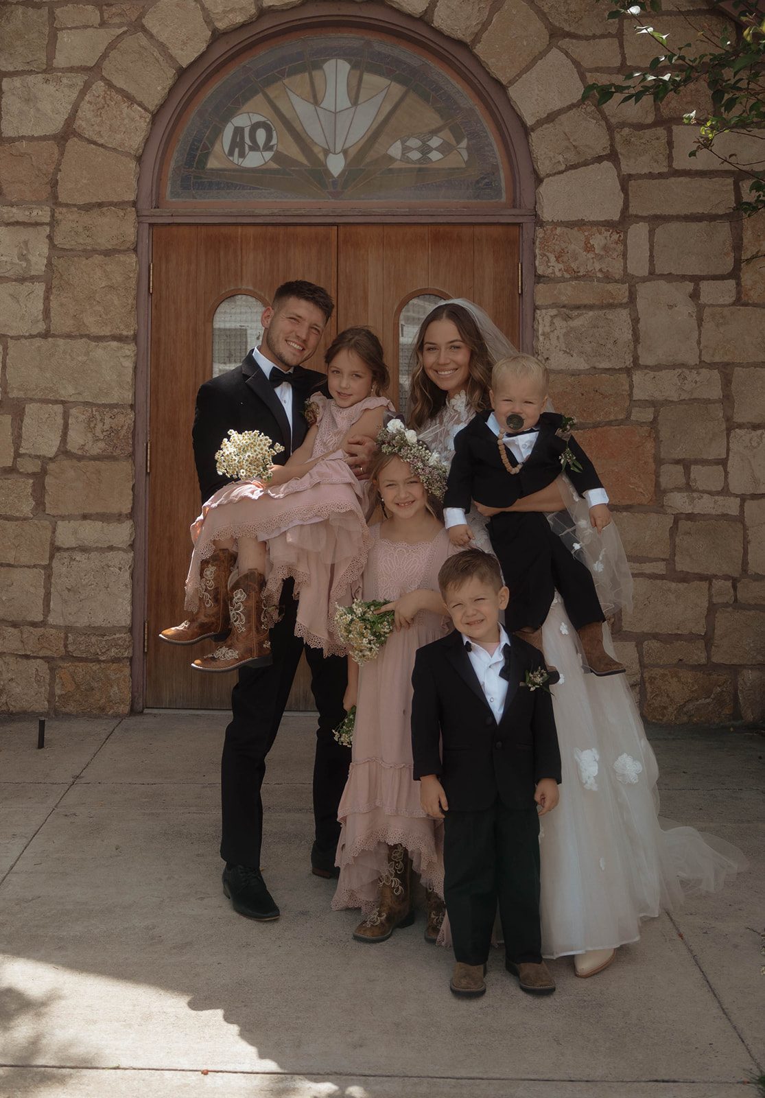 A group photo in front of a stone building shows a bride and groom with four children, two girls in pink dresses, and two boys in black suits. The bride is holding one child, and the groom holds another. | children at your wedding