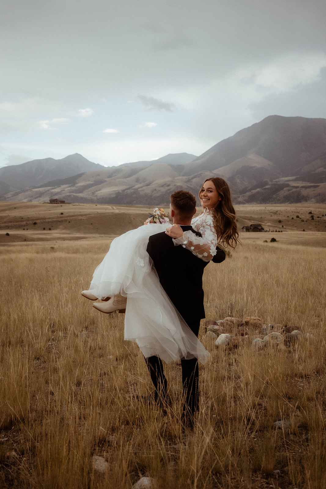 A bride and groom embrace in a field with mountains in the background. The bride is wearing a white wedding dress, and the groom is in a black suit 