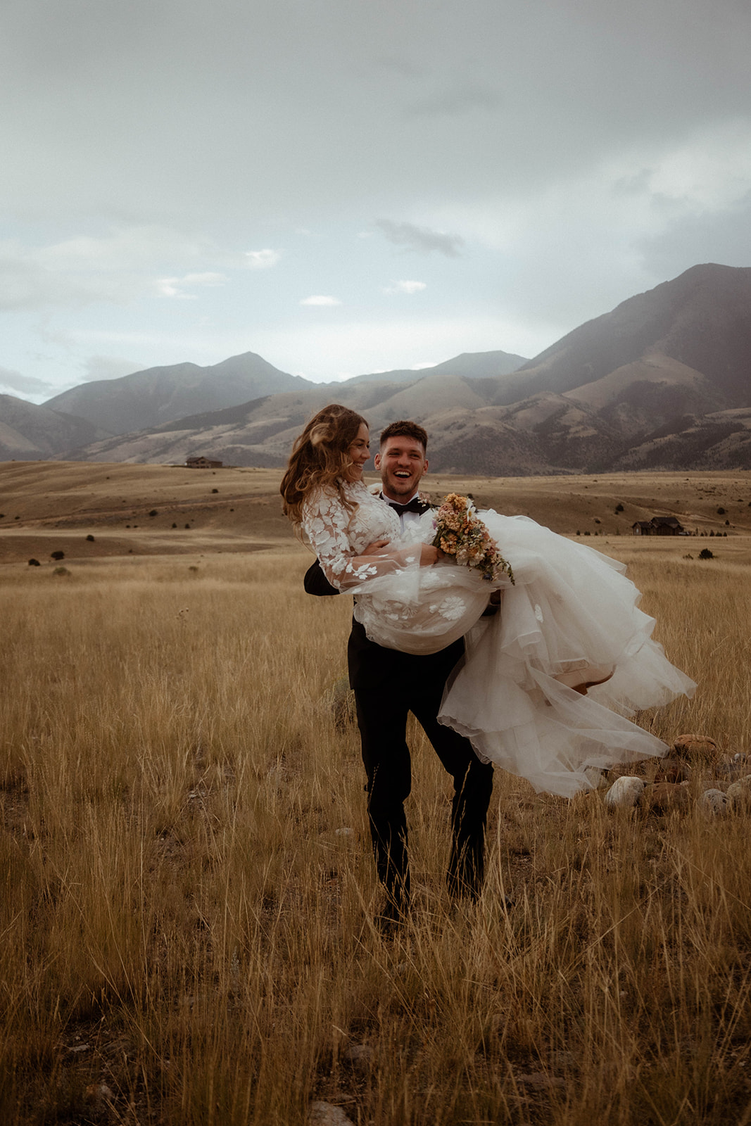 A bride and groom embrace in a field with mountains in the background. The bride is wearing a white wedding dress, and the groom is in a black suit for a wedding in Gardiner, Montana