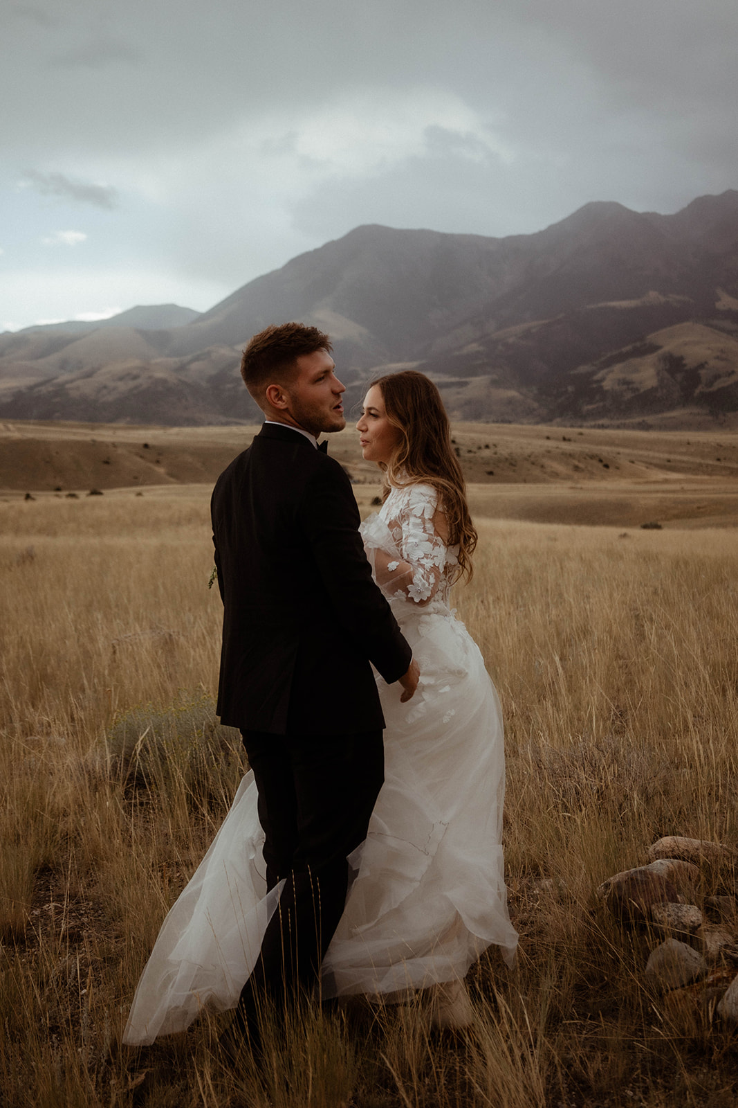 A bride and groom embrace in a field with mountains in the background. The bride is wearing a white wedding dress