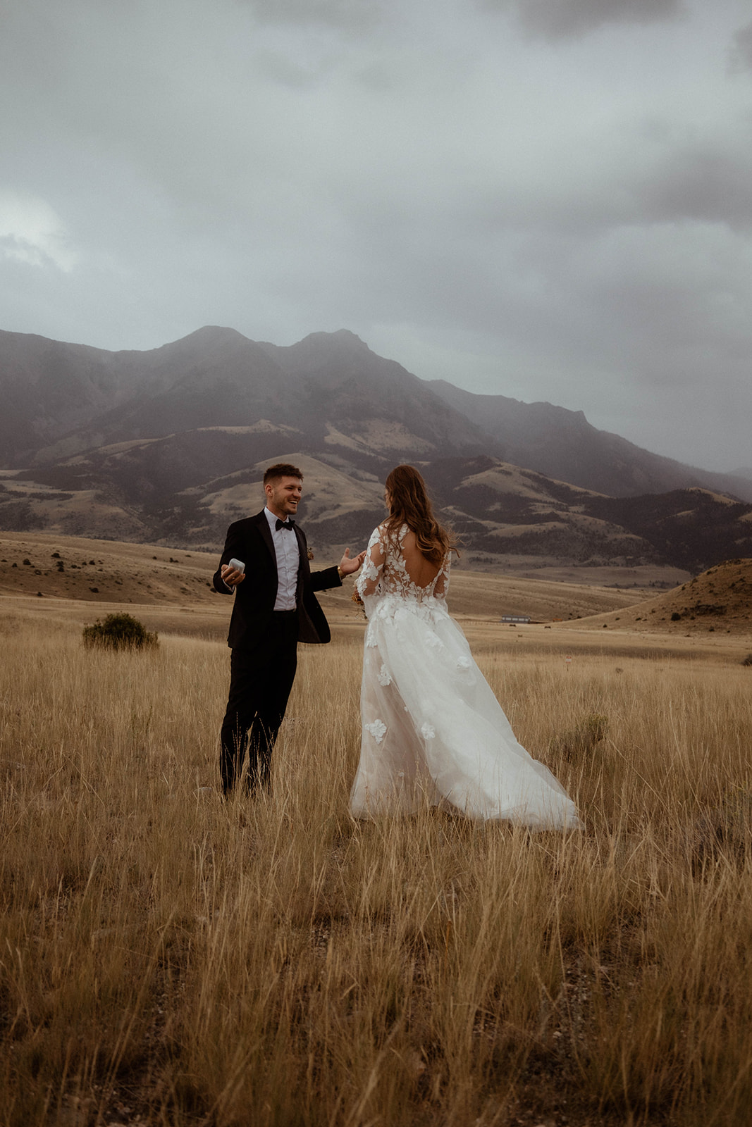 A bride and groom embrace in a field with mountains in the background. The bride is wearing a white wedding dress, and the groom is in a black suit for a wedding in Gardiner, Montana