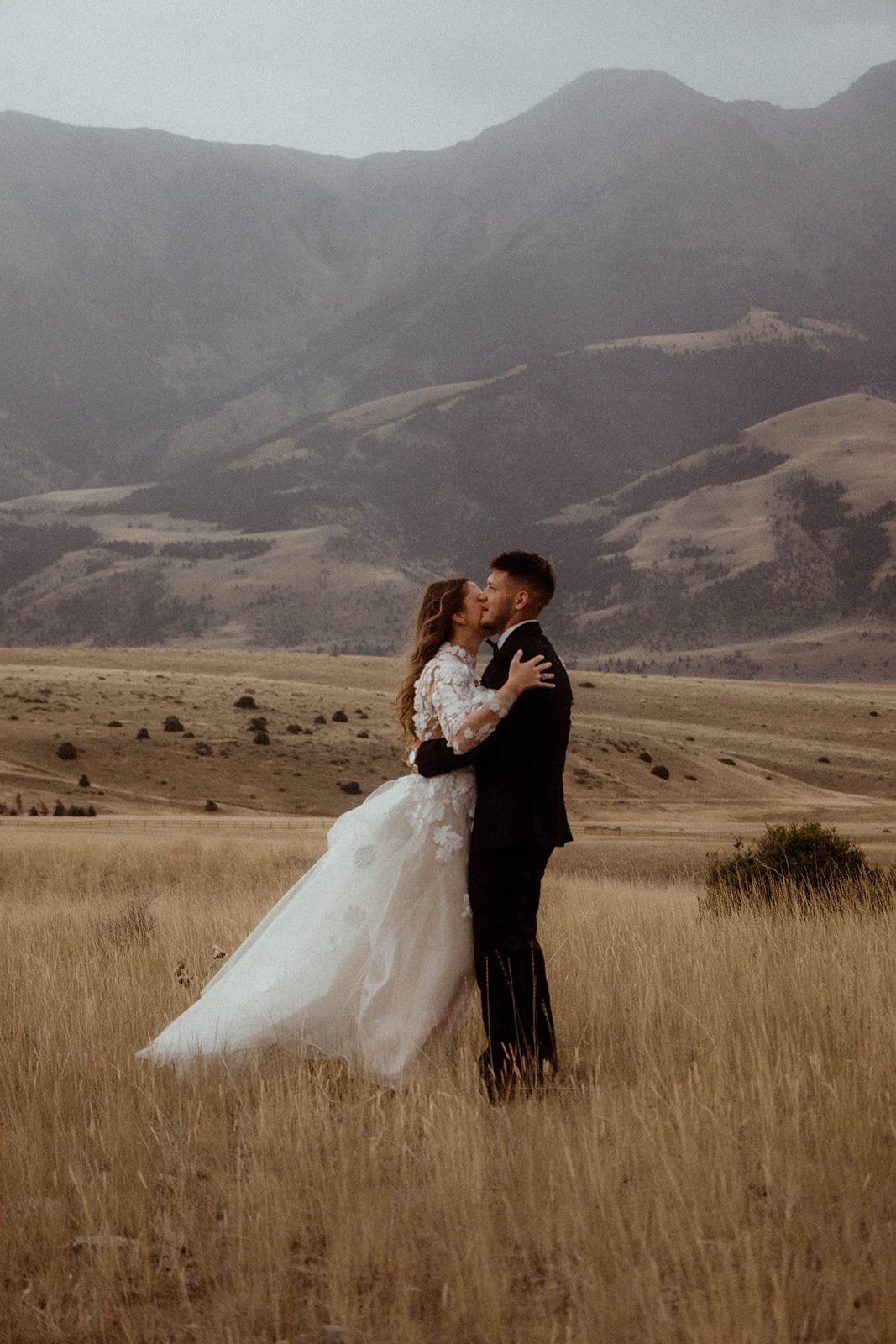 A bride and groom embrace in a field with mountains in the background. The bride is wearing a white wedding dress, and the groom is in a black suit 