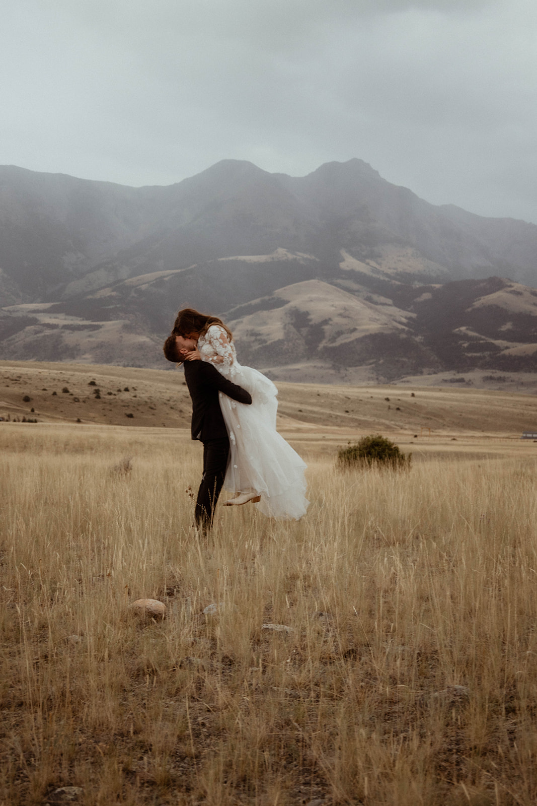 A bride and groom embrace in a field with mountains in the background. The bride is wearing a white wedding dress, and the groom is in a black suit 