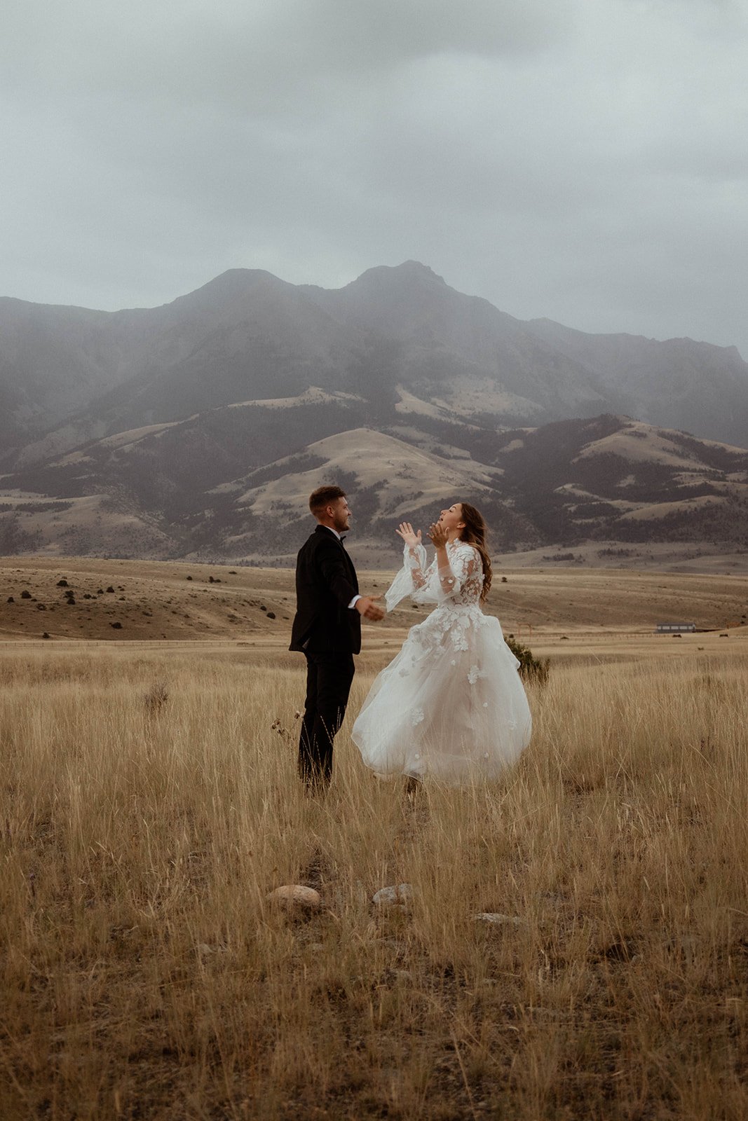 A bride and groom embrace in a field with mountains in the background. The bride is wearing a white wedding dress, and the groom is in a black suit 