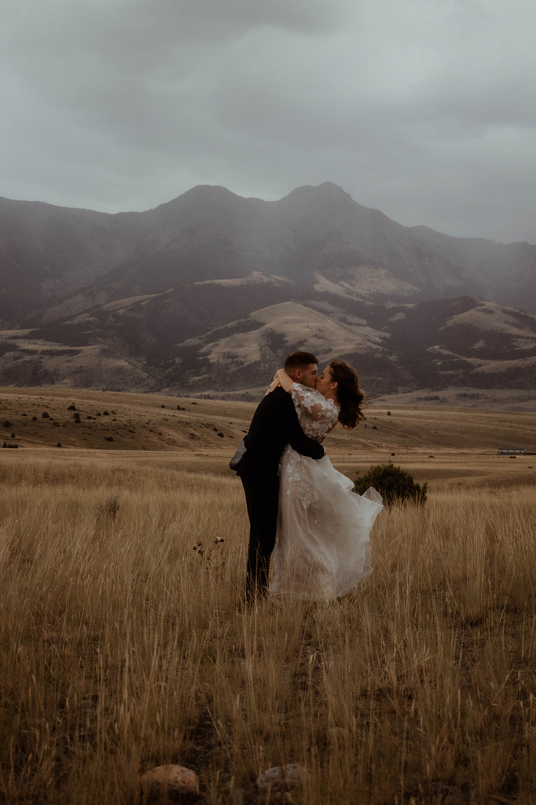 A bride and groom embrace in a field with mountains in the background. The bride is wearing a white wedding dress, and the groom is in a black suit