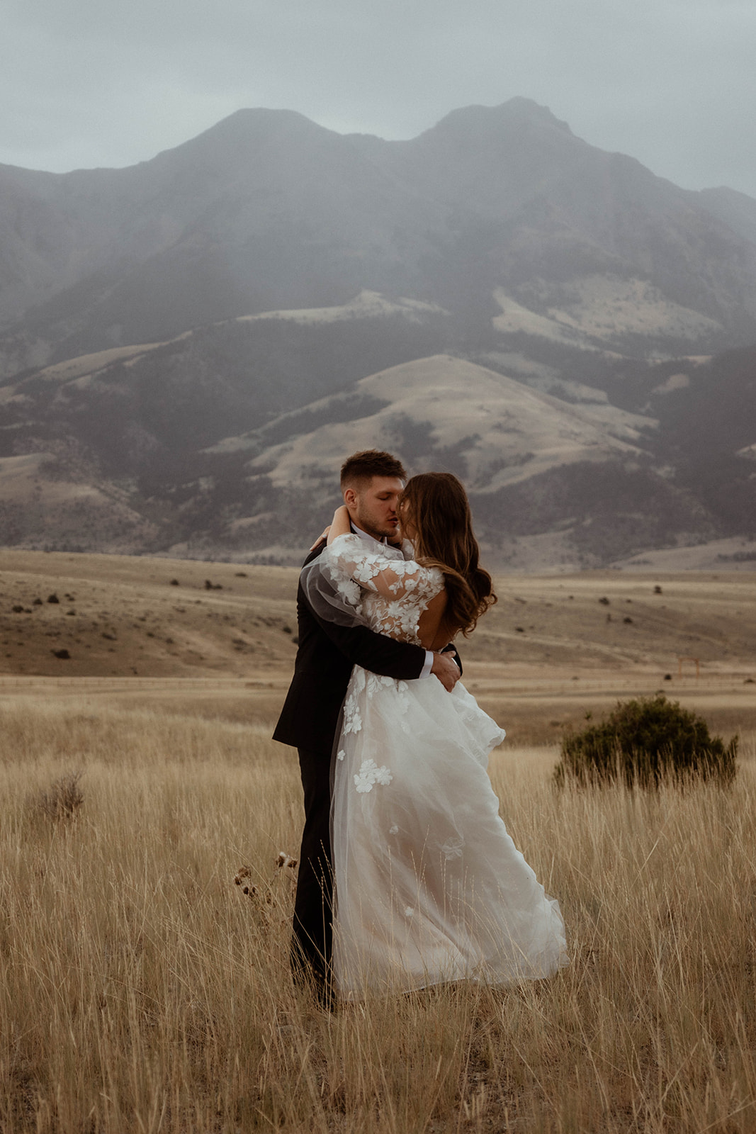 A bride and groom embrace in a field with mountains in the background. The bride is wearing a white wedding dress, and the groom is in a black suit