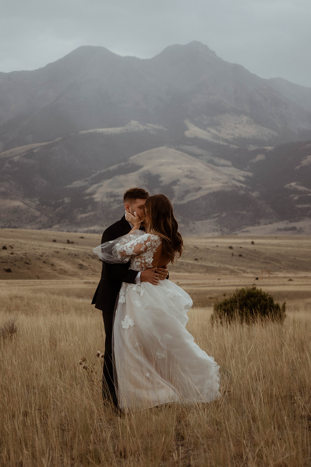 A bride and groom embrace in a field with mountains in the background. The bride is wearing a white wedding dress, and the groom is in a black suit 