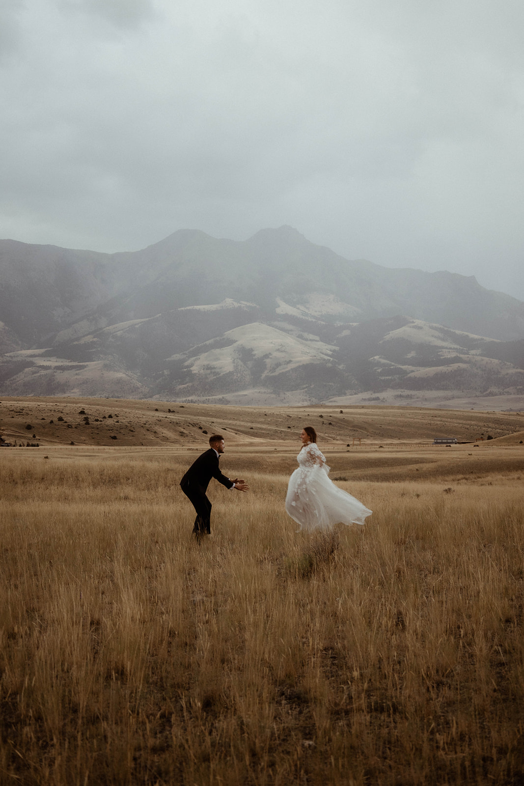 A bride and groom embrace in a field with mountains in the background. The bride is wearing a white wedding dress, and the groom is in a black suit 