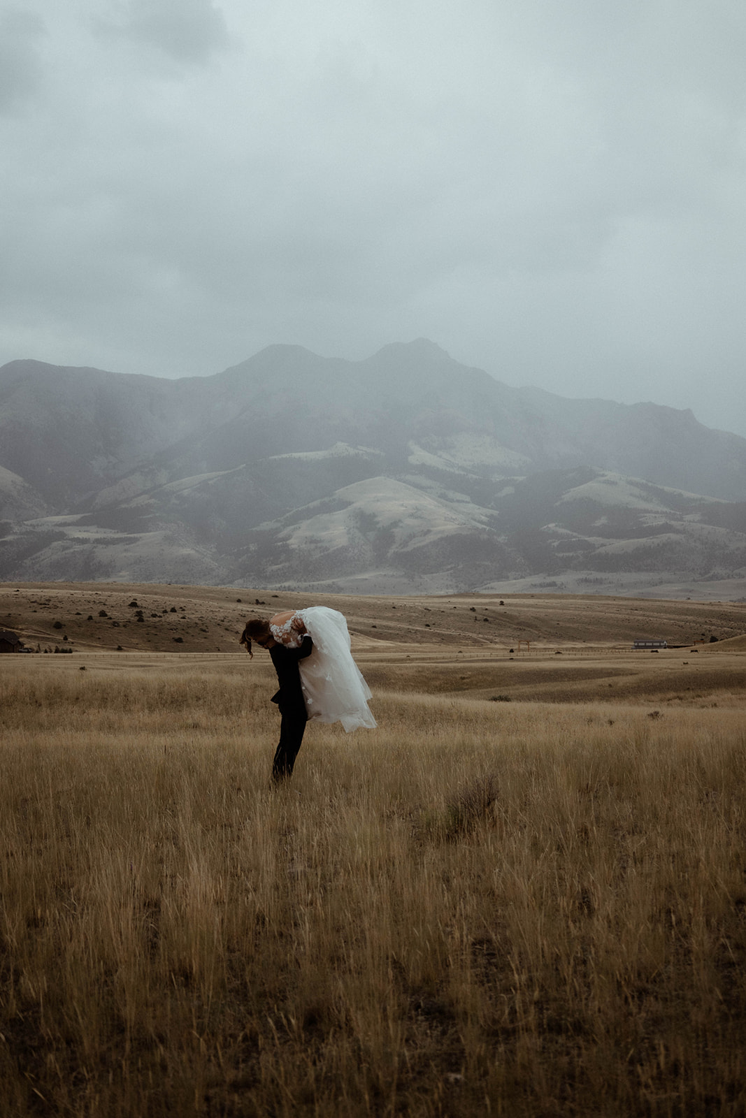 A bride and groom embrace in a field with mountains in the background. The bride is wearing a white wedding dress, and the groom is in a black suit 