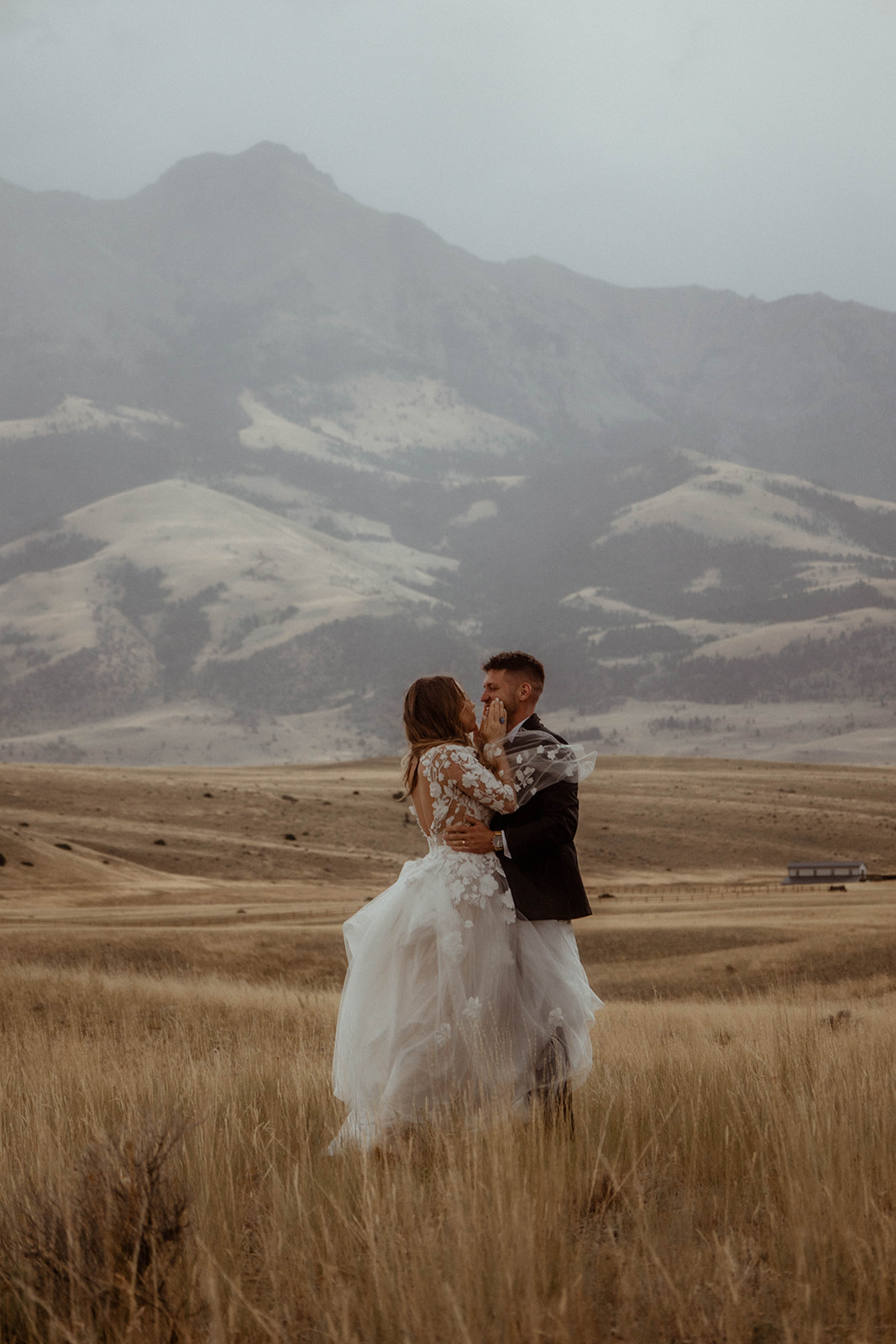 A bride and groom embrace in a field with mountains in the background. The bride is wearing a white wedding dress, and the groom is in a black suit 