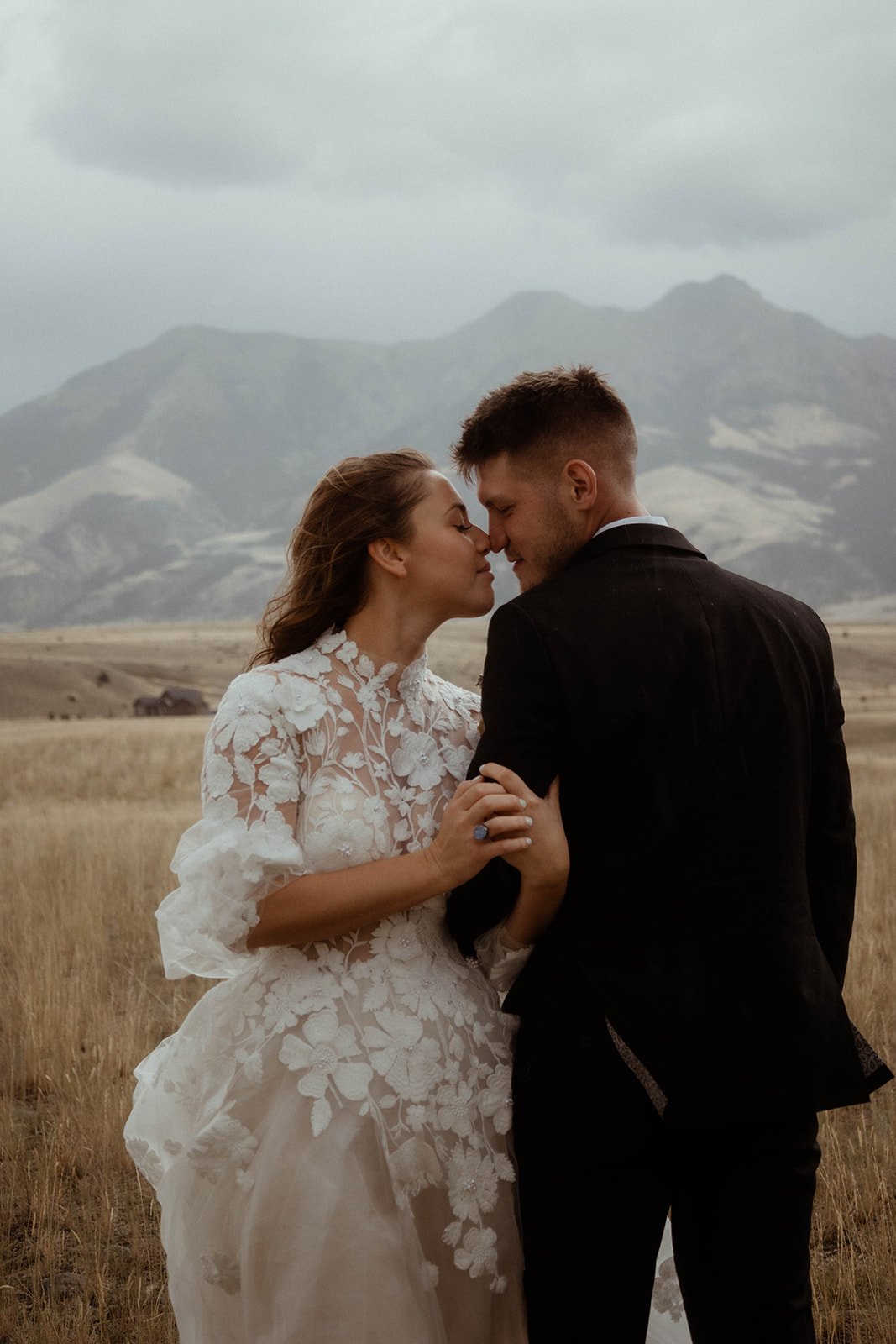 A bride and groom embrace in a field with mountains in the background. The bride is wearing a white dress, and the groom is in a black suit 