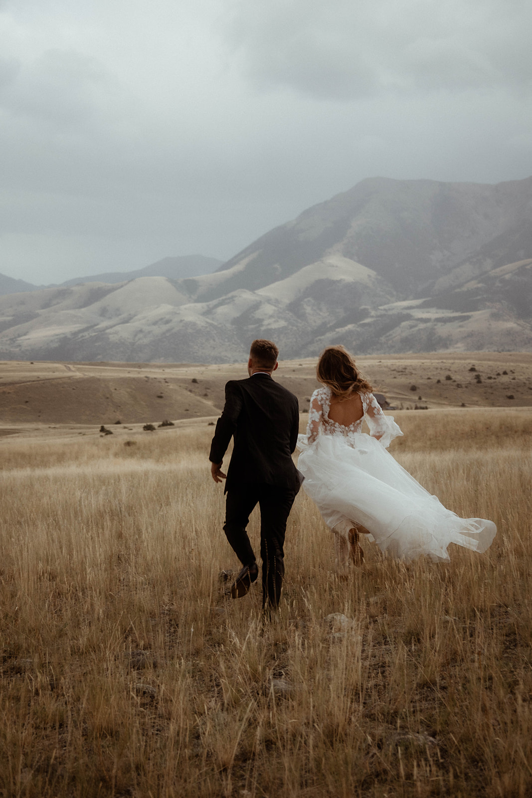 A bride and groom embrace in a field with mountains in the background. The bride is wearing a white  dress, and the groom is in a black suit