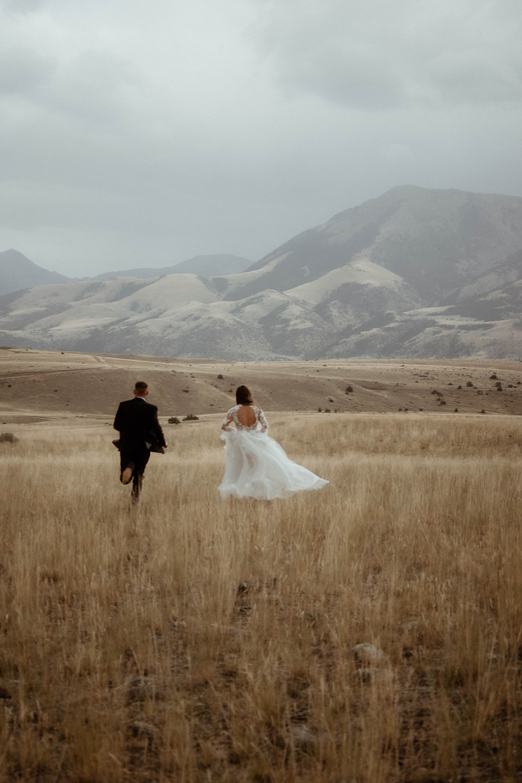 A bride and groom embrace in a field with mountains in the background. The bride is wearing a white  dress, and the groom is in a black suit 