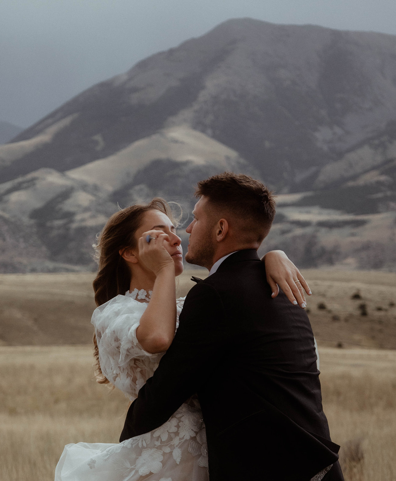 A bride and groom embrace in a field with mountains in the background. The bride is wearing a white wedding dress, and the groom is in a black suit for a wedding