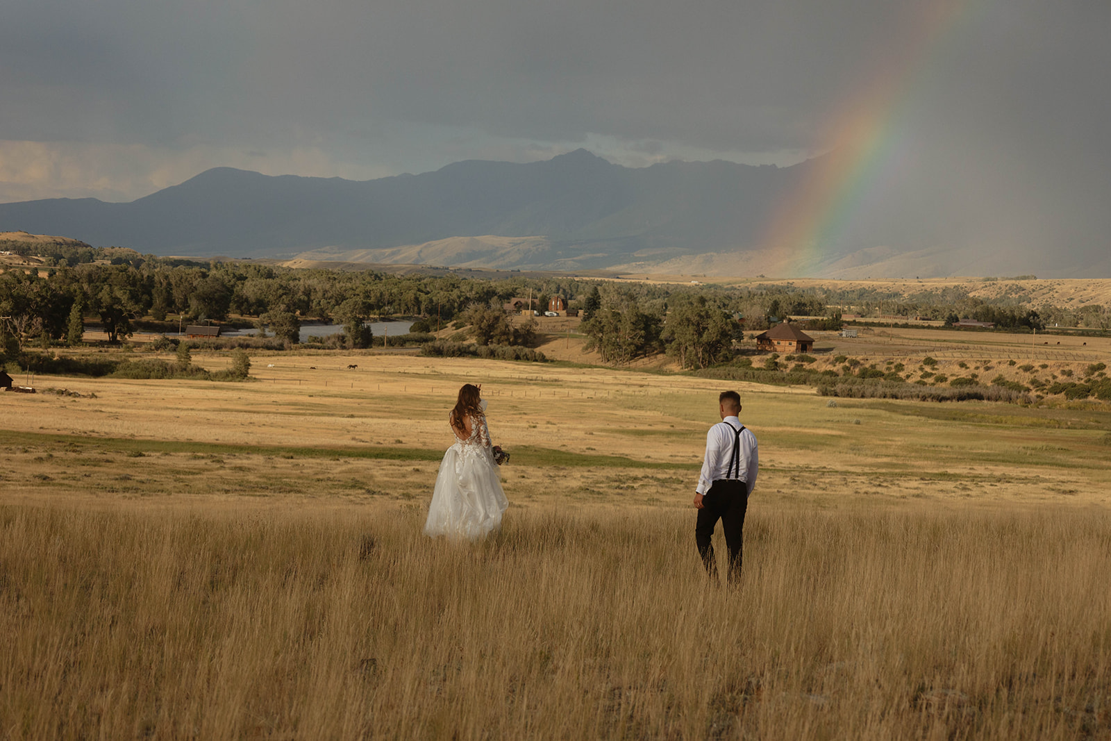 A bride and groom embrace in a field with mountains in the background. The bride is wearing a white wedding dress, and the groom is in a black suit for a wedding