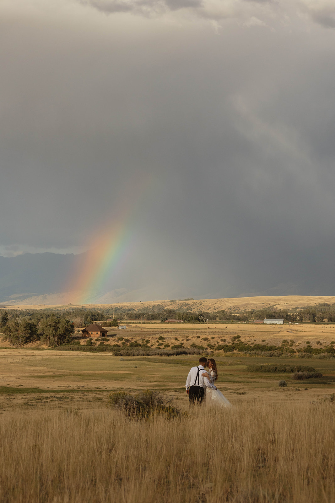 A couple in wedding attire stands in a field under a cloudy sky with a rainbow in the background.